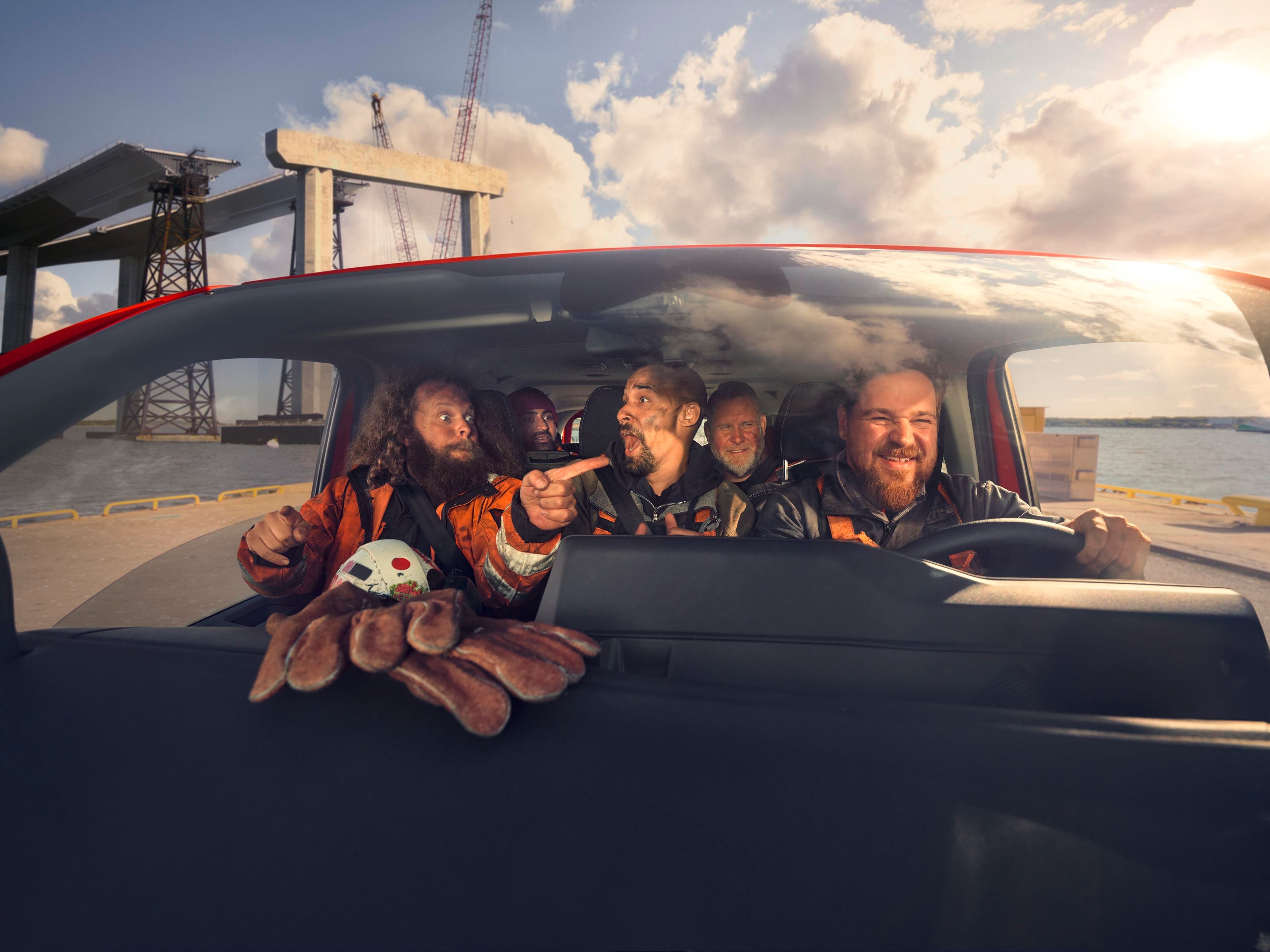 Mehrere Personen in Sicherheitskleidung sitzen in einem Auto und scheinen sich eine Baustelle mit Kranen und Stahlkonstruktionen anzusehen. Eine Person zeigt mit dem Finger in Richtung der Baustelle. Auf der Armaturenbrettablage liegt eine Spielzeugkrabbe.