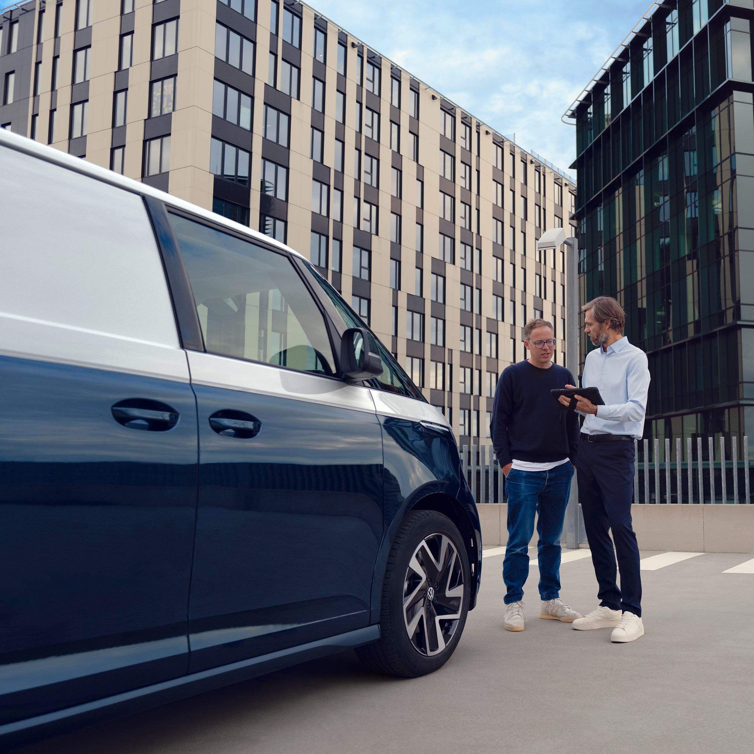 Zwei Personen stehen neben einem blauen Lieferwagen auf einem Parkplatz, umgeben von modernen Bürogebäuden. Eine Person zeigt der anderen etwas auf einem Tablet.