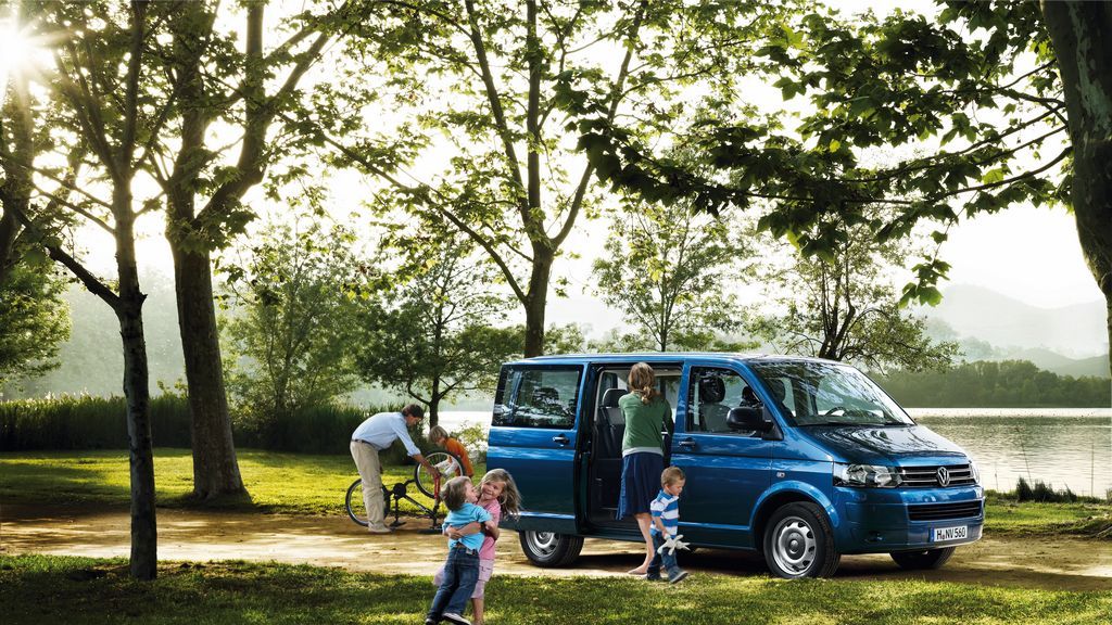 VW Kamper Bus blu qëndron përpara një liqeni. Fëmijët vrapojnë përreth. 